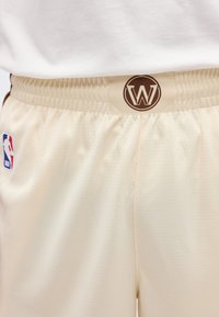 Μπεζ σορτς μπάσκετ με ελαστική μέση, κυκλικό λογότυπο "W" στο κέντρο και λογότυπο NBA στην αριστερή πλευρά.