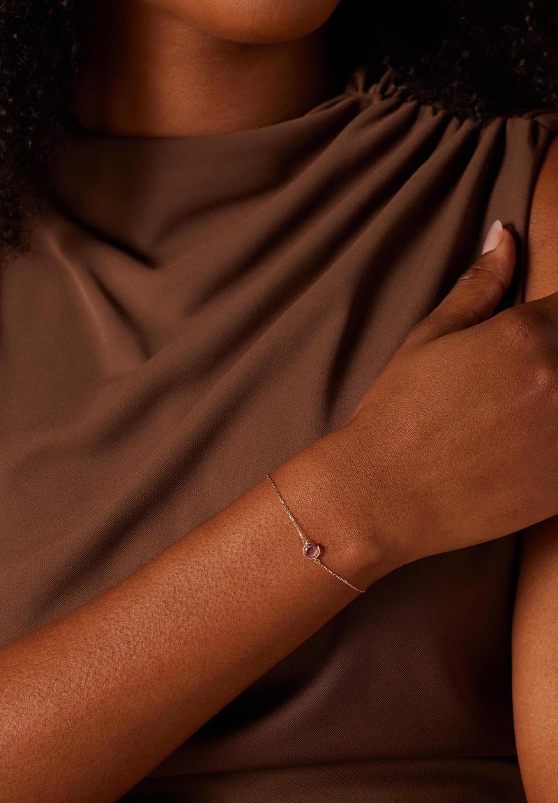 Zilveren schakelarmband met een roze edelsteen in het midden, gedragen om een bruin huidtype arm tegen een neutrale bruine stoffen achtergrond.
