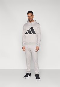 Ανοιχτό γκρι φούτερ με κουκούλα και μαύρο γεωμετρικό λογότυπο, σε συνδυασμό με ανοιχτό γκρι φόρμα. Λεπτομέρειες περιλαμβάνουν κουκούλα με κορδόνι και μαύρα παπούτσια.