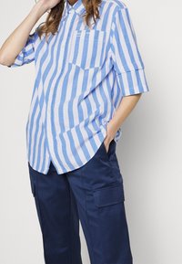 Camicia a maniche corte a righe blue e bianche con taschino sul petto, realizzata in tessuto leggero, indossata con pantaloni cargo blu navy.