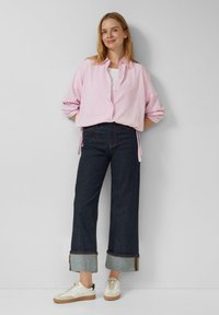 Kobieta stoi oparta o białą ścianę, ubrana w jasnoróżową koszulę na guziki, ciemne, szerokie jeansy z mankietami oraz białe trampki z gumowymi podeszwami.