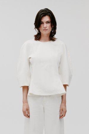 Femeie cu păr închis la culoare până la umeri, purtând cercei de argint în formă de cerc, top alb cu mâneci bufante și pantaloni albi cu picioare largi, pe un fundal simplu.