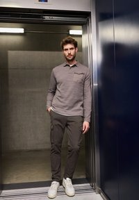 Grå långärmad polo med bröstficka, ihopparad med mörka cargo-byxor och sneakers, stående i en hiss. Betongbakgrund.