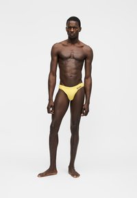 Športovo vyzerajúci muž stojaci bosý pred jednoliatym pozadím, oblečený v žltých plavkách s čiernym logom na ľavej strane.