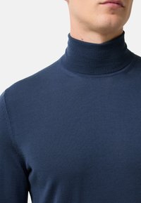Zbliżenie na osobę w gładkim granatowym swetrze z golfem, pokazującą szyję i górną część ciała na tle białym.