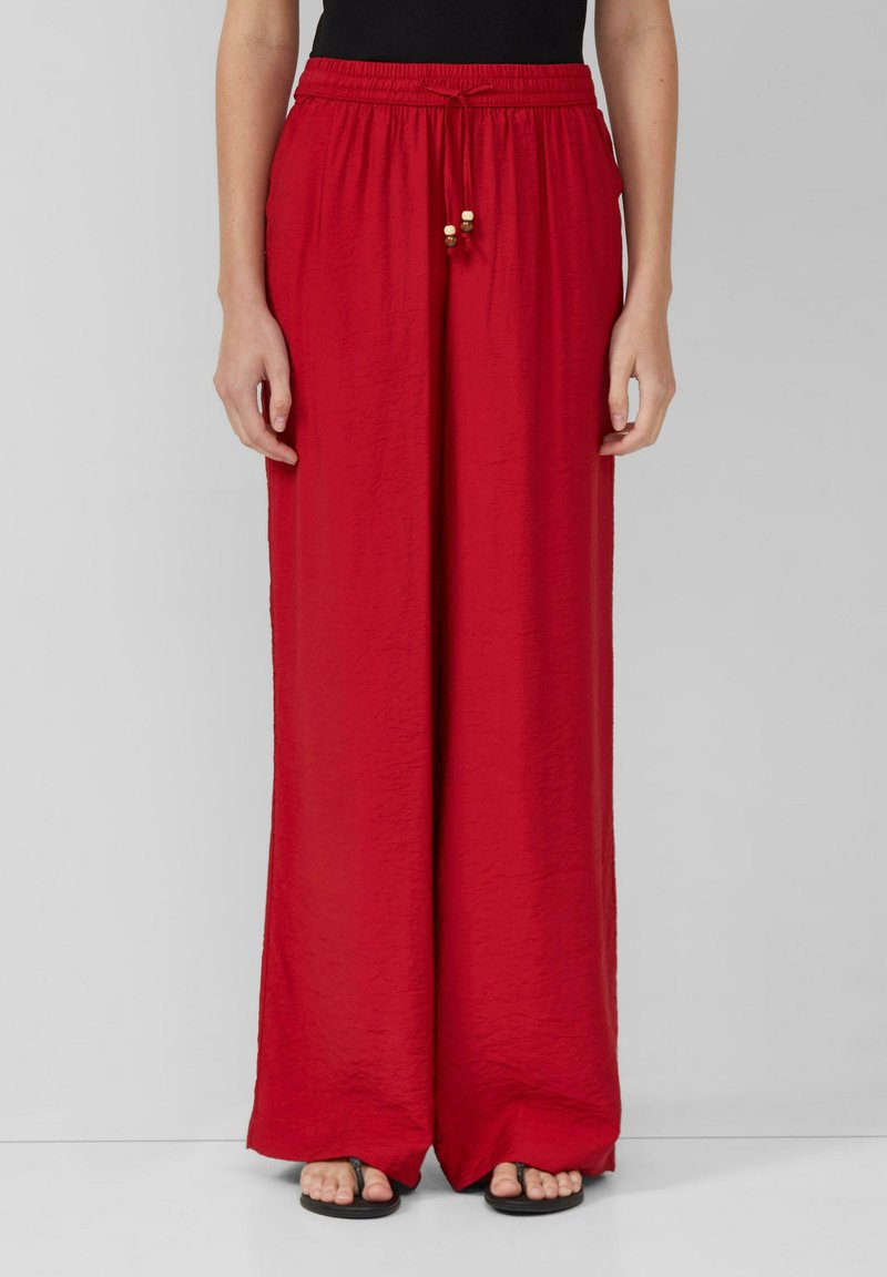 Pantalons larges rouges en tissu léger et texturé, avec une taille élastique et un cordon de serrage, ornés de perles en bois comme accents.