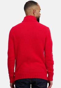 Pull rouge en tricot avec un col montant, doté d'un ourlet et de poignets côtelés, présenté de dos sur un fond uni.