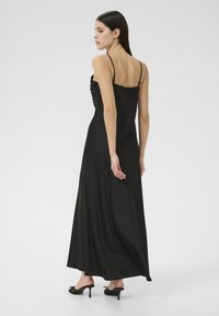 Žena s dugom tamnom kosom, obučena u crnu haljinu do poda s tankim naramenicama i crnim sandalama s visokim potpeticama, prikazana odostraga.