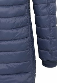 Section de veste matelassée bleu marine montrant des coutures horizontales et un poignet élastique sur la manche, fabriquée en tissu matelassé brillant.