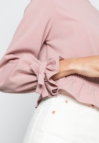 Pink blouse with ruffled sleeves and bow accents, featuring a gathered waistband. Paired with white pants, showcasing metal hardware details.