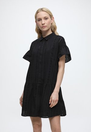 Žena s blond vlasmi nosí čierne šaty na gombíky s krátkymi rukávmi a rozšírenou sukňou, stojaca pred jednoduchým svetlým pozadím.