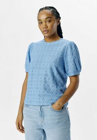 Ljusblå blus med puffärmar gjord av texturerad spets med diamantmönster och rund halsringning, tillsammans med ljusblå denimjeans.