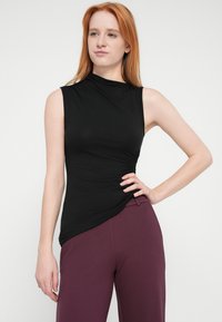 Schwarzes ärmelloses Oberteil mit hohem Ausschnitt und gerafftem Seitendetail, kombiniert mit bordeauxroten Hosen. Glatter Stoff mit figurbetonter Silhouette.