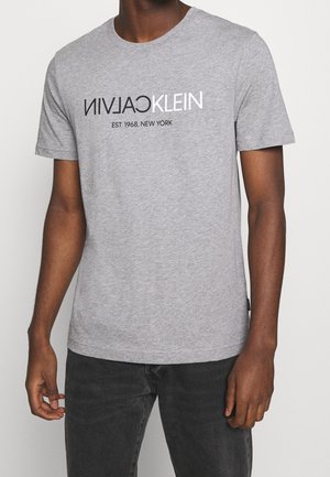 Homme portant un t-shirt gris à manches courtes avec l'inscription "CALVIN KLEIN EST. 1968, NEW YORK" en noir et blanc sur la poitrine.