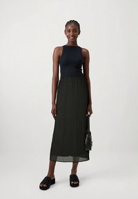 Črni brezrokavnik, pregibana midi krilo olivno zelene barve in črne platform sandale. Model drži majhno črno torbico. Nevtralna podlaga.