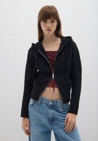 Felpa nera con zip realizzata in tessuto morbido, caratterizzata da un orlo a costine e un cappuccio con coulisse, abbinata a jeans blu chiaro a vita alta.