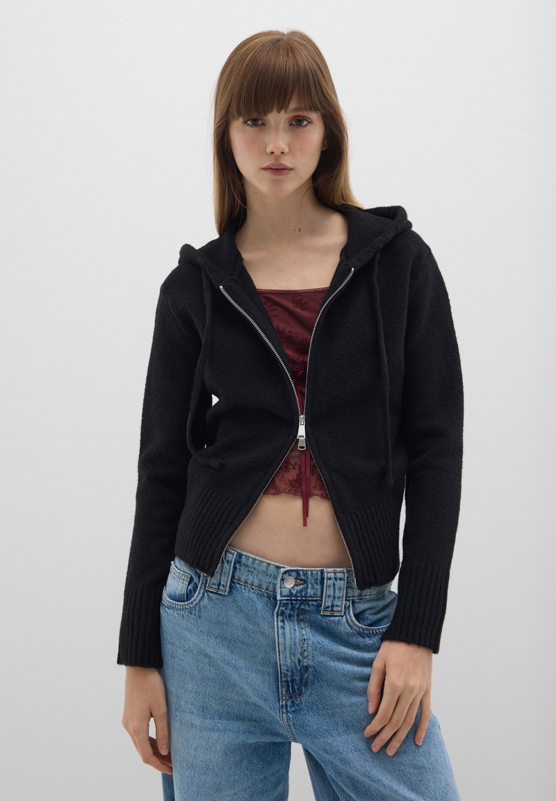 Felpa nera con zip realizzata in tessuto morbido, caratterizzata da un orlo a costine e un cappuccio con coulisse, abbinata a jeans blu chiaro a vita alta.