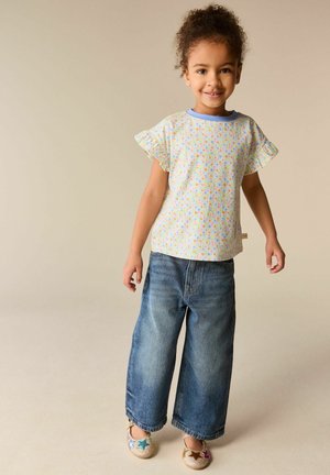 Mosolygó gyermek virágos rövid ujjú felsőt, bőszárú kék farmert és színes csillagmintás csillogó cipőt visel, bézs színű padlón áll.