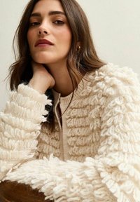 Maglione di colore crema con una trama a coste, caratterizzato da accenti soffici lungo le maniche e un colletto con bottoni. Materiale morbido e accogliente.