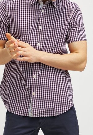 Man draagt een paars-wit geruit shirt met korte mouwen, houdt zijn handen samen, met marineblauwe broek tegen een effen lichte achtergrond.