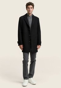 Musta villakangastakki, jossa on yksinkertainen muotoilu, napit, leveät kaulukset ja rento istuvuus, yhdistetty harmaaseen napilliseen paitaan ja harmaisiin housuihin.