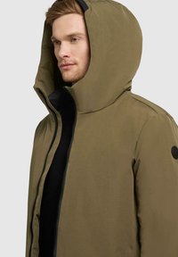 Olivenfarbene Kapuzenjacke mit hohem Kragen, Reißverschluss und schwarzen Akzenten. Gefertigt aus glattem, mattem Stoff mit minimalistischem Design.