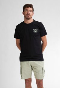 Črna bombažna majica s retro grafiko motocikla v beli barvi, kombinirana s svetlo zelenimi cargo kratkimi hlačami z stranskimi žepi.