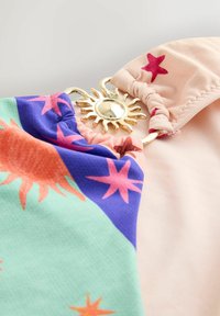 Két textilszakasz csillag- és napmintákkal, amelyeket egy arany, nap formájú gyűrű köt össze fehér háttéren.