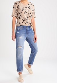 Camiseta beige con estampado de pájaros negros, combinada con jeans de mezclilla claros y desgastados y zapatillas color crema. Corte relajado y puños enrollados.