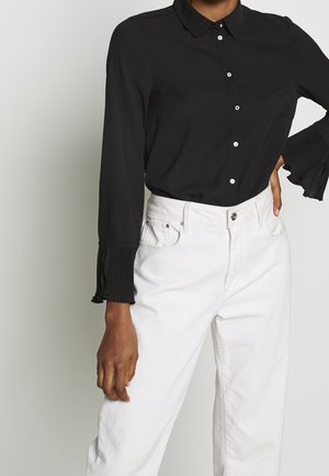 Person trägt eine schwarze Langarmbluse mit plissierten Glockenärmeln und weiße, hoch taillierte Jeans, Hand ruht auf Hüfte und Seite.