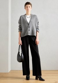 Cardigan de lã cinza com decote em V, frente com botões e ombros caídos, combinado com calças largas pretas e uma mala preta.