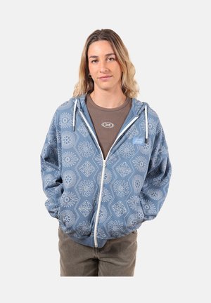 Jeune femme portant un sweat à capuche bleu zippé avec des motifs floraux blancs par-dessus un t-shirt marron, debout les mains dans les poches devant un fond uni.