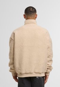 Jachetă din fleece bej cu guler înalt, croială supradimensionată și manșete ribbate. Material moale cu textură și talie netedă.