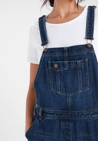Salopette en denim avec des bretelles réglables, une poche avant et des boutons latéraux. Présente une couleur bleu délavé et un tissu texturé.