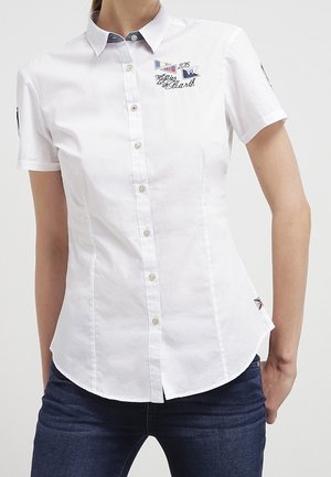 Chemise blanche à manches courtes avec boutons, avec des patchs brodés bleus et du texte, portée avec un jean bleu foncé sur une personne debout.