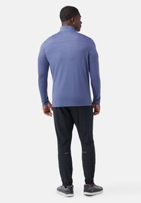 Maglietta sportiva a maniche lunghe, di un leggero color viola, con colletto alto, abbinata a pantaloni neri a tubo e sneakers grigie con texture.