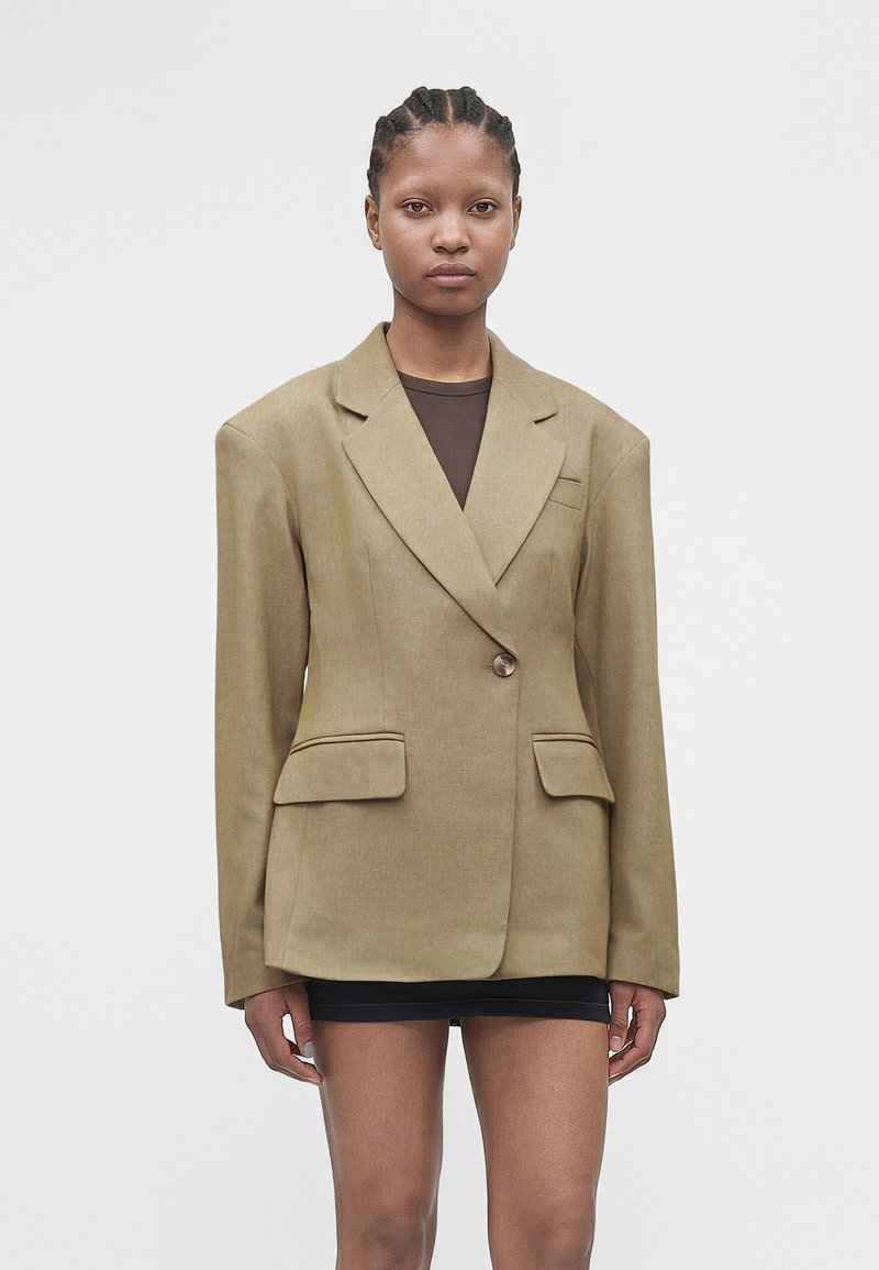 Γυναίκα με πλεξούδες, φορώντας ένα φαρδύ μπεζ σακάκι πάνω από ένα σκούρο τοπ και μια κοντή μαύρη φούστα, που στέκεται μπροστά σε ένα απλό φόντο.