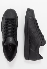 Ett par svarta lädersneakers från Adidas med snörning, skalmunddesign, och märkets signaturtre ränder på en vit bakgrund.