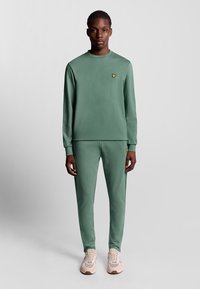 Felpa verde e pantaloni jogger coordinati in tessuto morbido, con un piccolo logo giallo sul petto. Scarpe da ginnastica di colore chiaro completano il look.