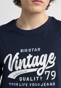 Naktinai mėlynos spalvos medvilnės marškiniai su dideliu, nusidėvėjusiu baltu grafiniu tekstu, kuriame užrašyta "Vintage 79 Quality Your Life Your Jeans."