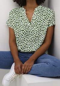 Green and white short-sleeved blouse featuring a leopard print, with a V-neckline and lightweight, textured fabric. Paired with blue jeans and sneakers.