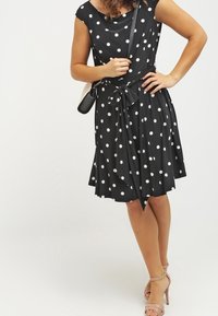 Mustapuku, jossa on valkoisia pilkkuja, istuva yläosa, levenevä helmasta ja sidottu solmu vyötäröllä. Yhdistettynä ihonvärisiin nilkkaketjullisiin korkokenkiin ja pieneen laukkuun.