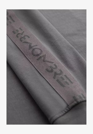 Tissu gris avec une texture douce, comportant une large bande avec "VON 32" imprimé en gris foncé. Le design est minimaliste et moderne.
