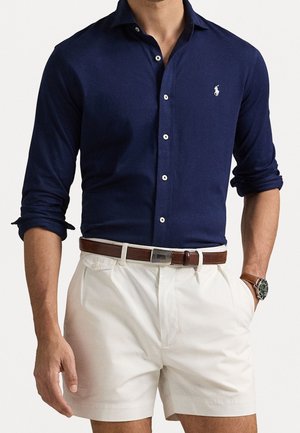 Man draagt een marineblauw overhemd met lange mouwen, opgerolde mouwen, witte korte broek, bruine riem en een polshorloge aan de linkerhand.