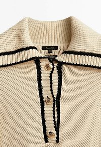Haut en coton beige tricoté avec une texture côtelée, détails rayés noirs le long du col et de la patte de boutonnage, avec des boutons en ton or.