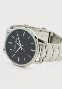 Ασημί ρολόι από ανοξείδωτο χάλυβα με υφασμένο μαύρο καντράν, ασημί δείκτες ωρών και μοντέρνα σχεδίαση βραχιολιού. Διαθέτει έντονο κορώνα.