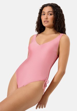 Mujer con cabello rizado oscuro que lleva un traje de baño entero rosa con escote en V y detalle de lazo lateral, sobre un fondo blanco liso.