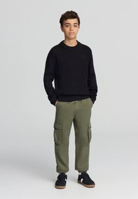 Czarny sweter z dzianiny o okrągłym dekolcie, w połączeniu z oliwkowymi spodniami cargo z bocznymi kieszeniami oraz czarnymi trampkami z brązowymi podeszwami.