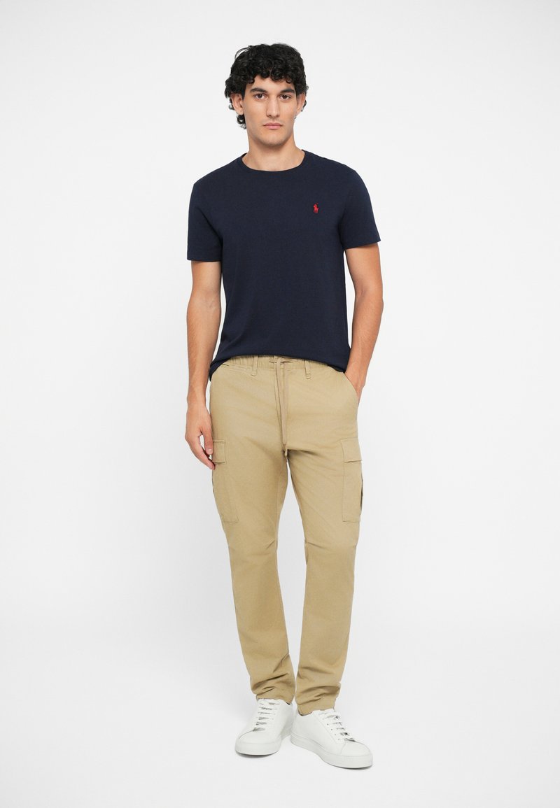 Marineblauwe crewneck t-shirt met een klein logo, gecombineerd met beige cargobroek met zijzakken, en witte sneakers.