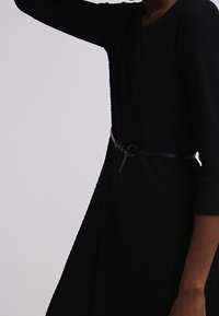 Vestido negro con textura, con un corpiño semi ajustado y falda acampanada, estilizado con un cinturón negro delgado en la cintura. Mangas de tres cuartos.